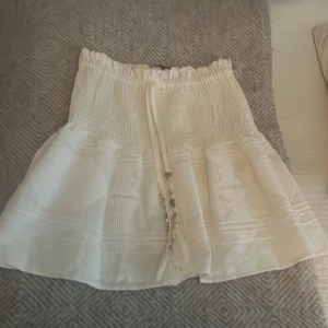 Vit kortkjol med broderade detaljer från Zara - Supersöt vit kortkjol från Zara med smockad midja och knytband med tofsar och små snäckor. Kjolen har broderade mönster och är perfekt för varma dagar. Lätt och luftig känsla, passar till många olika stilar.