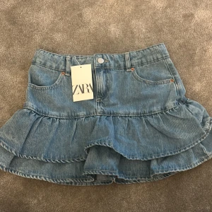 Blå volangkjol i jeans från Zara - Supersöt blå jeanskjol från Zara med volangdetaljer och klassisk femficksdesign. Kjolen har en kort längd och stängs med knapp och dragkedja framtill. Perfekt för dig som gillar denim och vill ha en trendig look.
