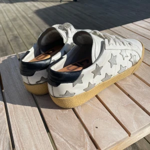 Saint Laurent skor ‼️intresse koll‼️ - Säljer ett par vita sneakers från saint Laurent  med silverfärgade stjärnor på sidorna och svart detalj vid hälen. Skorna har snörning och en beige gummisula med fiskbensmönster. Perfekta för dig som vill sticka ut med en unik och trendig look.