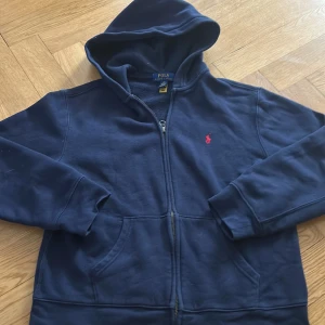 Mörkblå hoodie från Polo Ralph Lauren - Mörkblå hoodie med dragkedja från Polo Ralph Lauren. I princip oanvänd, mycket bra skick. Klassisk design med huva, fickor framtill och röd broderad logga på bröstet. Perfekt för en avslappnad stil. Nypris: 1100kr