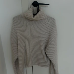 Beige ribbad polotröja - Säljer en beige ribbad polotröja med lång ärm och hög krage. Tröjan är stickad och har en avslappnad passform, perfekt för kyliga dagar. Enkel och stilren design som passar till det mesta.