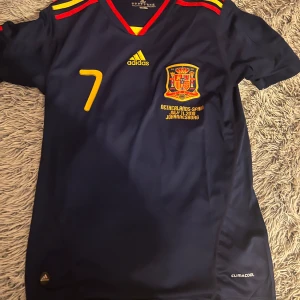 Spansk landslagströja David Villa 2010 - Mörkblå Spanien fotbollströja från Adidas med nummer 7 och David Villa på ryggen. Tröjan har gula och röda detaljer, landslagsemblem och text om VM-finalen 2010. ClimaCool-material och korta ärmar.
