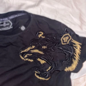 Svart t-shirt med tigerbroderi från Philipp Plein - Cool svart t-shirt från Philipp Plein med ett stort broderat tigerhuvud i gult och svart på framsidan. Rund halsringning och korta ärmar. Märkesdetaljer i nacken och på bröstet. Perfekt för dig som vill sticka ut.