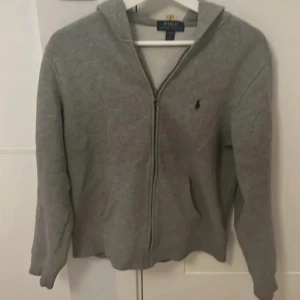 Grå hoodie från Polo Ralph Lauren - Säljer en klassisk grå hoodie från Polo Ralph Lauren med dragkedja och huva. Tröjan har långa ärmar, två fickor framtill och den ikoniska broderade loggan på bröstet. Skulle säga att den passar en person med storlek S 