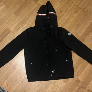 Monclear windbreaker - Svart vindjacka från Moncler med huva och dragkedja. Jackan har vita och röda ränder på huvan samt Moncler-logga på ärmen. Justerbar snörning i huvan och två fickor framtill med tryckknappar. Perfekt för höstdagar och regniga sommardagar. Storlek L. 