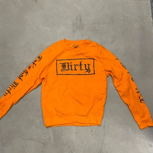 Orange sweatshirt från Dirty - Säljer en orange sweatshirt från Dirty med svart tryck på bröstet och längs ärmarna. Tröjan har rund hals och långa ärmar. Perfekt för dig som vill sticka ut med en färgstark och streetinspirerad stil.
