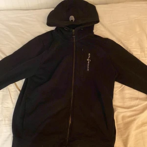 Svart hoodiejacka från Sail Racing - Svart hoodiejacka från Sail Racing med dragkedja framtill och huva. Jackan har en enkel design med logga på bröstet och är tillverkad i mjukt material som känns skönt mot huden. Perfekt för kyliga dagar när du vill vara både bekväm och snygg.