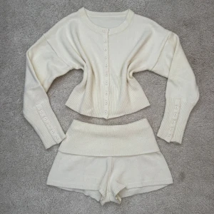 Vit stickad kofta och shorts set - Säljer ett krämvitt stickat set med croppad kofta och matchande shorts. Koftan har långa ärmar, rund halsringning och knappar framtill. Perfekt för en mysig och chill stil. 