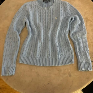 Stickad tröja Ralph Lauren - Säljer en klassisk ljusblå kabelstickad tröja från Ralph Lauren. Tröjan har rund halsringning, långa ärmar och är tillverkad i 100% bomull. Liten broderad logga på bröstet. Perfekt för lager-på-lager-stil. (Märkt L barn men passar mig som jag S)