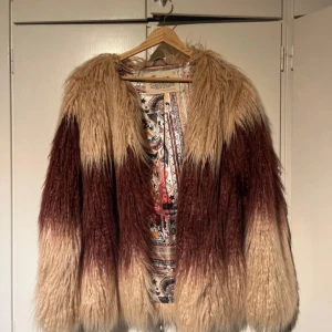 Beige och vinröd fuskpälsjacka från Odd Molly - Säljer en fluffig och statement beige och vinröd fuskpälsjacka från Odd Molly. Jackan har lång luggig päls, öppen framtill men har knappar och ett färgglatt paisleymönstrat innerfoder. 