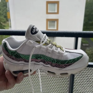 Nike Air Max 95 med gröna och rosa detaljer - Nike Air Max 95 sneakers i vitt med gröna och rosa ormskinnsmönstrade detaljer längs sidorna. Skon har snörning, synlig Air Max-dämpning i sulan och en sportig look. Perfekt för dig som vill sticka ut med färg och mönster.