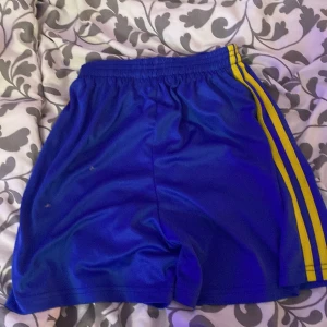 Blå sportshorts med gula ränder - Säljer ett par blå sportshorts med två gula ränder längs sidorna. Shortsen har resår i midjan och är tillverkade i 100% polyester, vilket gör dem lätta och bekväma för träning eller sportaktiviteter.