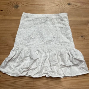 Vit volangkjol - Säljer en söt vit kjol med volang nertill. Kjolen har en smockad midja som ger en bekväm passform och en luftig känsla. Perfekt att styla med en enkel topp för en fräsch look.