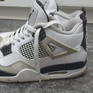 Nike Air Jordans 4 Retro White/Black - Säljer ett par klassiska Nike Air Jordans 4 Retro i vitt med svarta och grå detaljer. Skorna har nätpaneler på sidorna, snörning och synlig luftdämpning i sulan. Perfekta för dig som gillar streetwear och sneakers med ikonisk design.