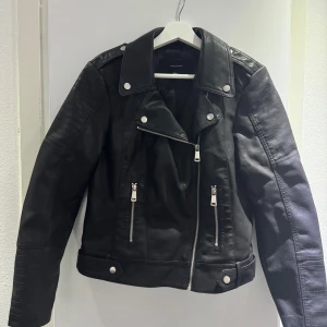 Svart skinnjacka från Vero Moda - Snygg svart skinnjacka från Vero Moda med silvriga dragkedjor och knappar. Jackan har klassisk bikerstil med bred krage och detaljerade sömmar på ärmarna. Perfekt för en cool och stilren look.