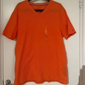 Orange t-shirt med v-ringning från Tommy Hilfiger - Snygg orange t-shirt från Tommy Hilfiger med v-ringad hals. Klassisk passform och korta ärmar, perfekt för en avslappnad stil. Liten logga på ärmen och tillverkad i mjuk bomull.