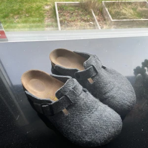 Grå Birkenstock tofflor i filt - Säljer ett par gråa Birkenstock tofflor i filtmaterial med justerbart spänne över foten. Skorna har en klassisk rund tå och platt sula, perfekta för att glida runt hemma eller på jobbet. Nypris 1850kr🤩