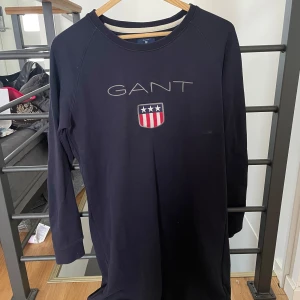 Svart långärmad klänning från GANT - Svart långärmad klänning från GANT med rund halsringning och broderad logotyp samt sköld med stjärnor och ränder framtill. Enkel och stilren design, perfekt för en avslappnad look.