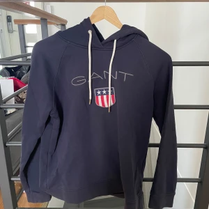 Mörkblå hoodie från GANT - Säljer en mörkblå hoodie från GANT med broderad logga och sköld på bröstet. Tröjan har huva med vita snören och långa ärmar. Perfekt för en avslappnad och stilren look.