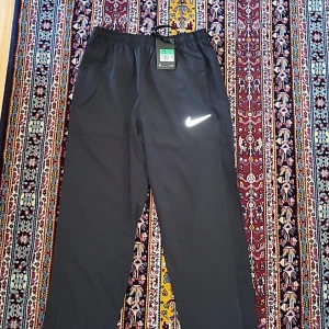 Svarta träningsbyxor från Nike Dri-Fit - Svarta Nike Dri-Fit träningsbyxor med resår i midjan och dragsko. Byxorna har två sidofickor och en vit Nike-logga på låret. Lätta och bekväma för löpning eller gym. Modellen är rak och materialet är syntet.