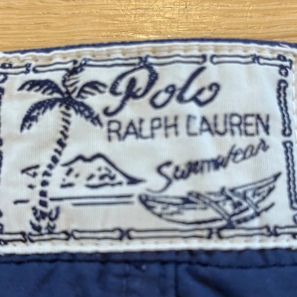 Snygga marinblå badshorts från Polo Ralph Lauren med röd broderad logga på benet och röd snörning i midjan. Klassisk modell med normal passform, perfekt för sommaren. Tillverkade i polyester för snabb torkning. Storlek: M (10-12). Hör av dig vid frågor!. Muu.