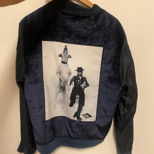 Mörkblå jacka, Limitato - Säljer en mörkblå bomberjacka med sammetsliknande material och svarta ärmar. På ryggen finns ett stort konstnärligt tryck i svartvitt med en person och en hund. Jackan har dragkedja framtill och ribbade muddar.