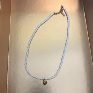 Vitt pärlhalsband med snäckberlock - Säljer ett halsband med små vita pärlor och en liten snäckformad berlock i guld. Halsbandet har justerbart spänne i silverfärg. Perfekt accessoar för en somrig look!