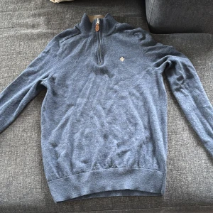 Blå merinoulltröja med halv dragkedja från Morris - Säljer en blå långärmad tröja från Morris i 100% merinoull. Tröjan har halv dragkedja vid halsen och en liten broderad logga på bröstet. Perfekt för dig som gillar klassisk och stilren design.