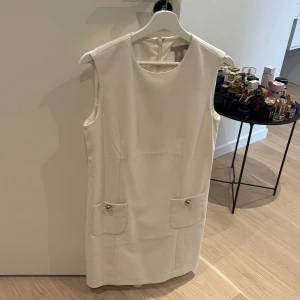 Vit ärmlös klänning med fickor från H&M - Säljer en stilren vit ärmlös klänning från H&M i storlek S. Klänningen har två fickor framtill med dekorativa guldfärgade knappar och en diskret dragkedja bak. Perfekt för en fräsch och klassisk look.