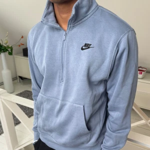 Ljusblå halvzip sweatshirt från Nike - Säljer en ljusblå sweatshirt från Nike med halv dragkedja och hög krage. Tröjan har en stor magficka och Nike-logga broderad på bröstet. Perfekt för en avslappnad och sportig stil.