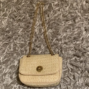 Beige axelväska med kedja - Säljer en beige axelväska i flätat material med guldfärgad kedjerem och snyggt vridlås framtill. Perfekt storlek för det viktigaste och ger en trendig touch till din outfit. Den är från H&M