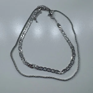 Två silverfärgade halsband - Säljer två snygga halsband i silverfärg. Det ena har en bredare, platt länkdesign medan det andra är tunnare med tvinnad kedja. Perfekta att bära tillsammans eller var för sig för en stilren look.