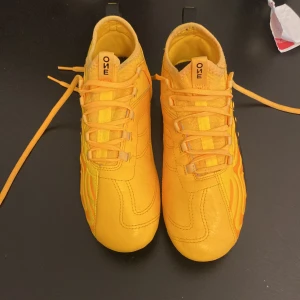 Gula fotbollsskor från Puma - Säljer ett par gula fotbollsskor från Puma med svarta detaljer och snörning. Skorna har rund tå och platt sula med dobbar för bra grepp på planen. Perfekta för dig som vill sticka ut på fotbollsplanen!