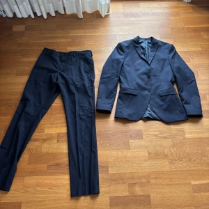 Mörkblå kostym med kavaj och byxor - Säljer en stilren mörkblå kostym med matchande kavaj och byxor. Kavajen har tre knappar framtill och klassiska fickor. Perfekt för formella tillfällen eller fest. Snygg och tidlös design.