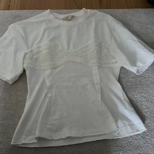 Vit t-shirt med spetsdetalj från H&M - Vit t-shirt från H&M med unik spetsdetalj över bröstet som ger en korsettliknande look. T-shirten har rund halsringning och korta ärmar. Perfekt för dig som vill ha en basic t-shirt med en twist.