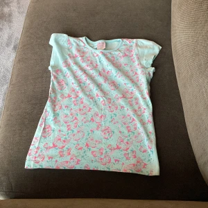 Ljusblå t-shirt med rosa mönster - Säljer en ljusblå t-shirt i 100% bomull med korta ärmar och ett gulligt rosa mönster över hela framsidan. Perfekt för dig som gillar färgglada och lekfulla plagg!