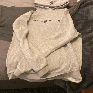 Grå hoodie från Sail Racing - Säljer en ljusgrå hoodie från Sail Racing med svart logga och tryck på bröstet samt på huvan. Tröjan har en klassisk känguruficka framtill och ribbade muddar vid ärmslut och nederkant. Perfekt för en avslappnad stil.