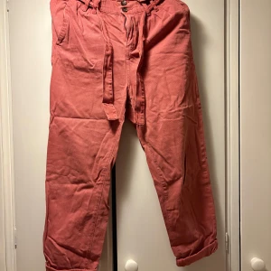 Rosa byxor med knytband från Denim - Säljer ett par rosa byxor från Denim med knytband i midjan och uppvikta benslut. Byxorna har fickor fram och bak, samt stängs med knappar och dragkedja. Perfekta för dig som gillar en avslappnad men snygg stil.