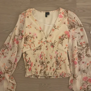 Blommig blus med volang från Vero Moda - Säljer en romantisk blommig blus från Vero Moda i cremefärgad ton med rosa och bruna blommor. Blusen har v-ringning, långa transparenta ärmar och smockad midja med små knappar framtill. Perfekt för vår och sommar!