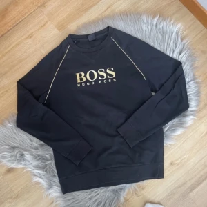 Svart sweatshirt från Hugo Boss - Säljer en svart sweatshirt från Hugo Boss med guldfärgad logga på bröstet och dekorativa sömmar längs axlarna. Tröjan har rund halsringning och långa ärmar. Perfekt för en stilren och avslappnad look.