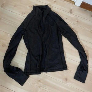 Svart träningsjacka med dragkedja - Säljer en svart, figurnära träningsjacka med hög krage och dragkedja framtill. Jackan har långa ärmar och är tillverkad i ett stretchigt syntetmaterial som passar perfekt till träning eller som ett extra lager.