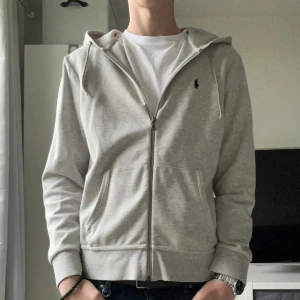 Ralph Lauren Zip - Säljer en stilren grå hoodie från Polo Ralph Lauren med dragkedja och huva. Tröjan har två fickor framtill och en liten broderad logga på bröstet. Perfekt för en avslappnad look.