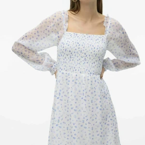 Vit blommig långärmad klänning med smock - Supersöt vit klänning med små blå blommor och smockad överdel. Långa, transparenta ärmar med resår vid handlederna och fyrkantig urringning. Perfekt för vår och sommar, ger en romantisk och luftig känsla.