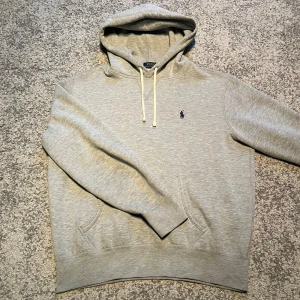 Ralph Lauren hoodie  - Säljer en stilren grå hoodie från Polo Ralph Lauren med klassisk broderad logga på bröstet. Tröjan har huva med vita snören och en stor magficka. Perfekt för en avslappnad och snygg look.