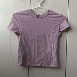 Ljuslila t-shirt från H&M - En basic ljuslila t-shirt från H&M med rund halsringning och korta ärmar. Perfekt att matcha med jeans eller kjol för en enkel och fräsch look. Tillverkad i mjuk bomull för skön komfort.