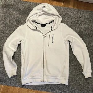 Vit hoodie från Sail Racing med dragkedja - Vit hoodie från Sail Racing med hel dragkedja framtill och huva. Jackan har en bröstficka med dragkedja och broderad logga på bröstet. Mjuk insida och ribbade muddar vid ärmslut och nederkant. Nypris 1000 en liten fläck kan man se på bild 4. Skickar snabbt 💨 