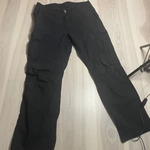 Svarta cargobyxor från Clockhouse - Svarta cargobyxor från Clockhouse med flera fickor på sidorna och bak. Byxorna har normal passform och stängs med knapp och dragkedja. Perfekta för en avslappnad stil och har en cool detalj på knappen framtill. OBS!! byxorna är i st 34/36 i ”manstorlek” och inte i ”kvinnorstorlek”. 