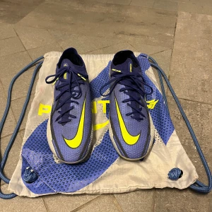 Nike Phantom GT2 Elite fotbollsskor blå/gul - Säljer ett par Nike Phantom GT2 Elite fotbollsskor i blått med gula detaljer och snörning. Skorna har en smal passform och är tillverkade i syntetmaterial för bästa grepp och kontroll på planen. Perfekta för dig som vill sticka ut på fotbollsplanen! Nästan aldrig använda så jag var skadad när jag köpte dom. Har en liten defekt men inget som märks, ena plastbiten på snöret är av. Man kan även köpa ny sån. Originalpris: 2899kr. Mitt pris: 1300kr. Pris går att diskutera 