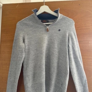 Grå halvzip tröja från Morris - Snygg grå tröja från Morris i 100% merinoull med halv dragkedja och diskret broderad logga på bröstet. Tröjan har ribbade muddar och hög krage, perfekt för lager-på-lager-stil.