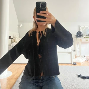 Svart tröja med knytning - Säljer en snygg svart tröja med knytning framtill från Ginatricot. Den har långa och breda ärmar och en stilren design som passar perfekt till både vardag och fest. Tröjan är i bra skick och ger en chic look. Den är helt ny och därav priset!